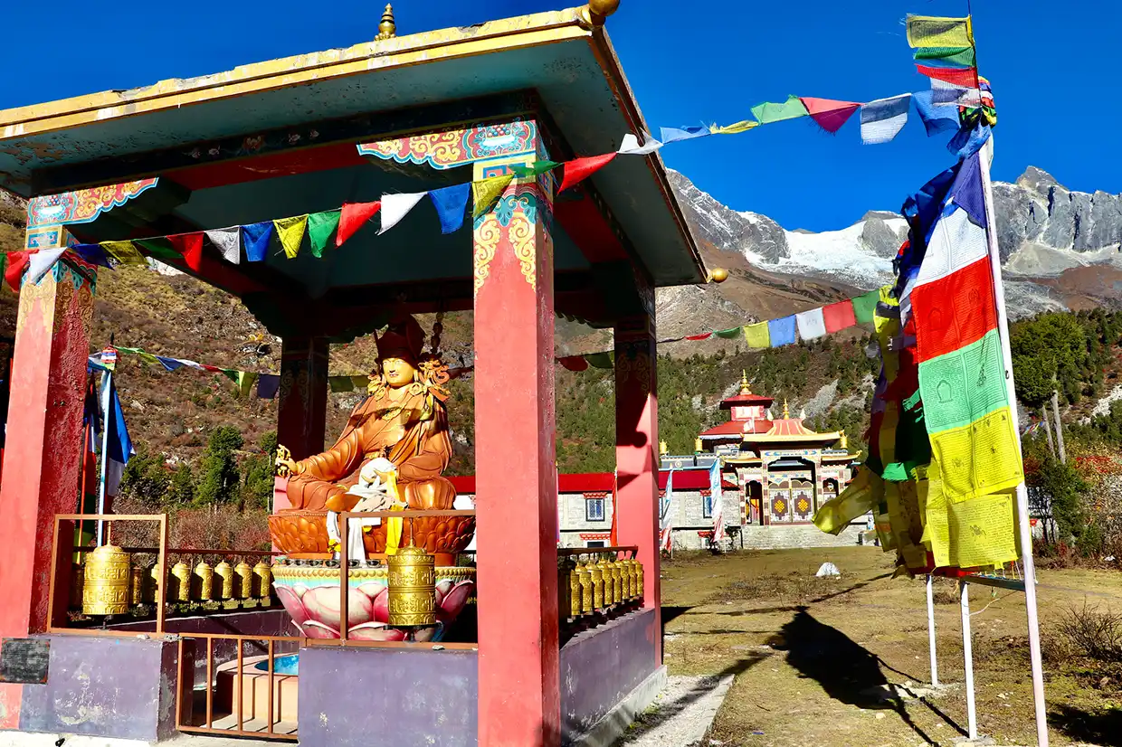 A golden Buddhist statue sitting inside an open shrine decorated with prayer flags, featuring a mountain peak background on the Manaslu Tsum Valley Trek.