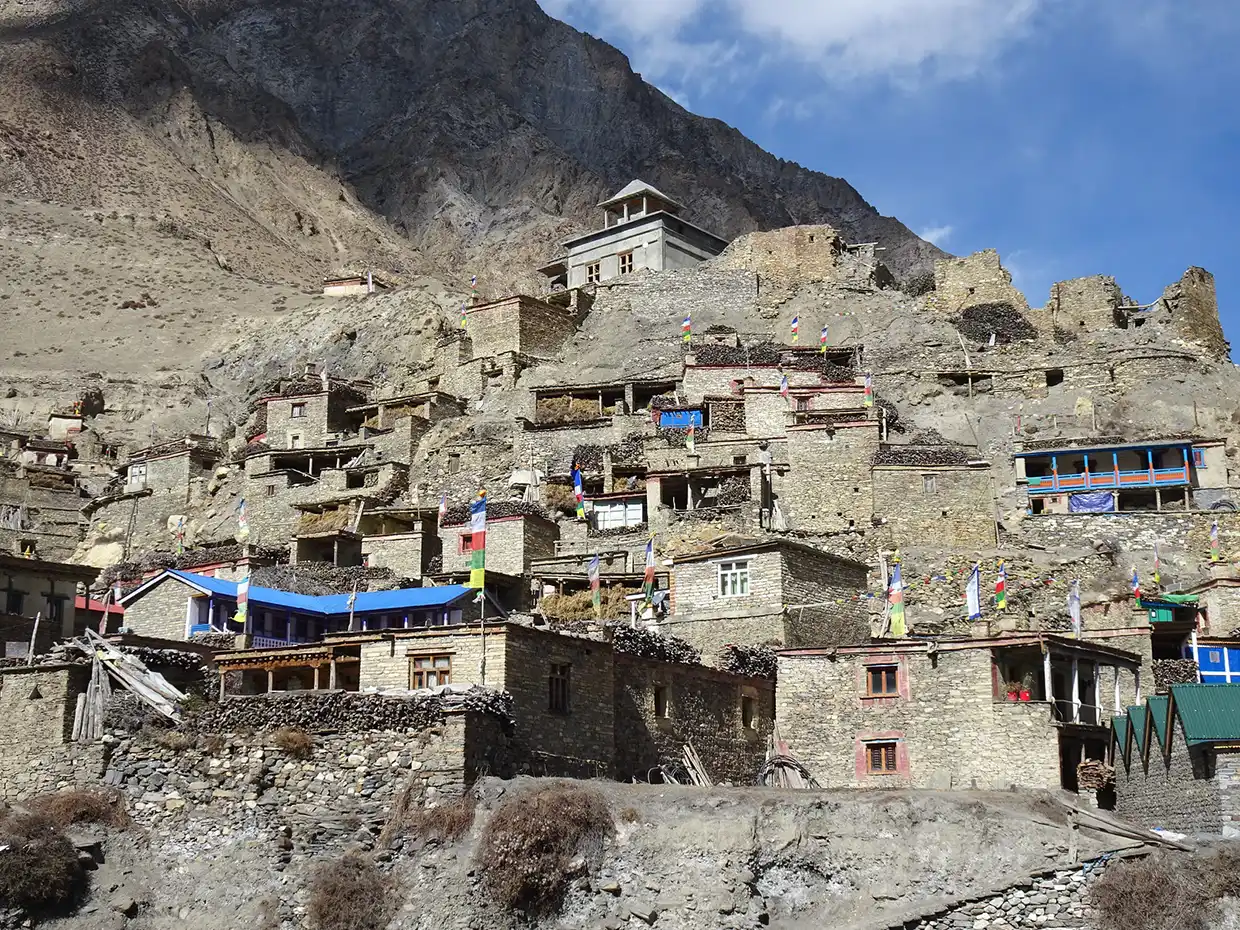 Traditional stone houses with colorful prayer flags crowd a steep hillside below towering mountain peaks during the scenic Nar Phu Valley Trek.