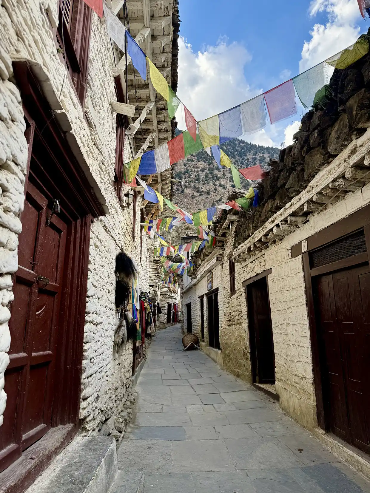 Colorful prayer flags hang across a stone paved alley with white traditional buildings during an Upper Mustang Jeep Tour in Nepal.