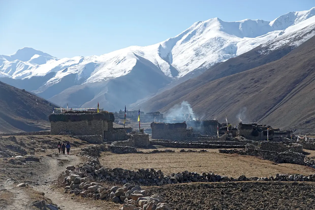 Hikers walk along a dirt path toward a traditional stone village with smoking chimneys, set against giant snow-capped mountains on the Upper Dolpo Trek.