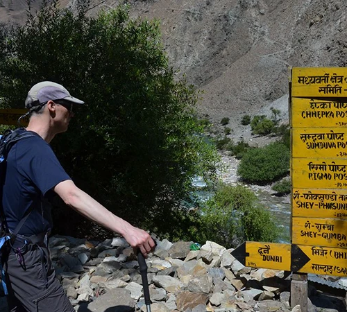 A hiker with a backpack and trekking pole stands on a rocky trail next to a yellow wooden signpost marking routes on the Upper Dolpo Trek.