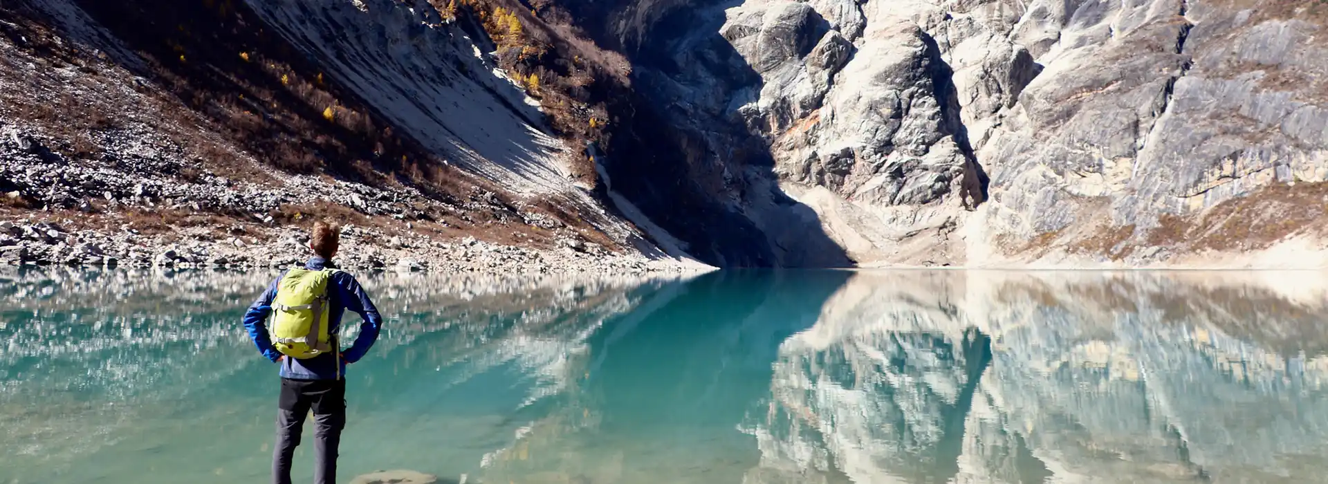 A hiker with a backpack stands at the edge of a still turquoise lake reflecting steep mountain walls along the Manaslu Tsum Valley Trek.