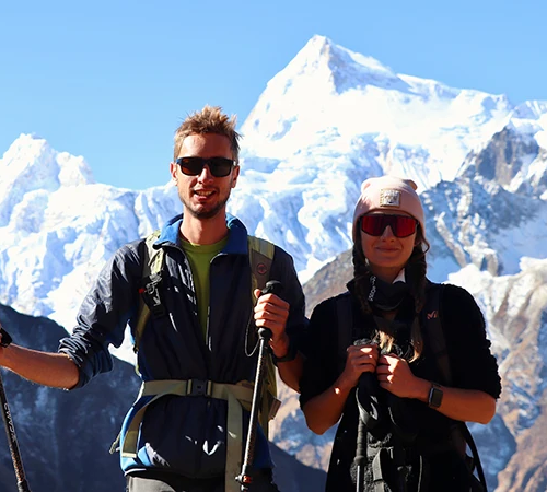 Two trekkers wearing sunglasses and carrying trekking poles smile in front of a massive, snow-covered mountain peak along the Manaslu Tsum Valley Trek route.
