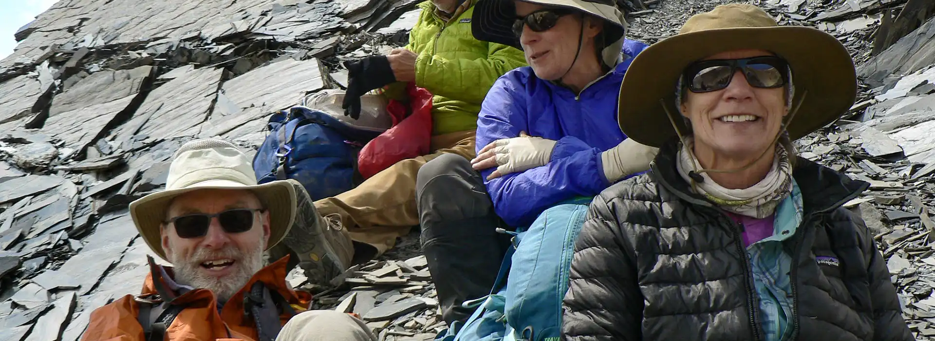 Four smiling hikers wearing sun hats and sunglasses take a rest break on a steep slope of sharp, layered slate rock during their Upper Dolpo Trek.