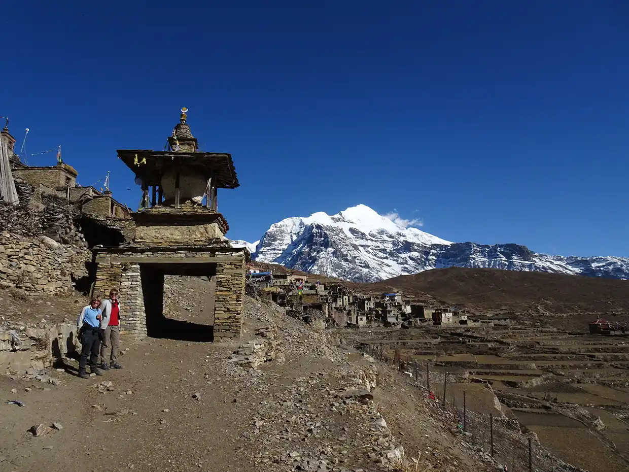 Two hikers stand next to a historic stone chorten gate near a mountain village with a snow-capped peak in the background during a Nar Phu Valley Trek.