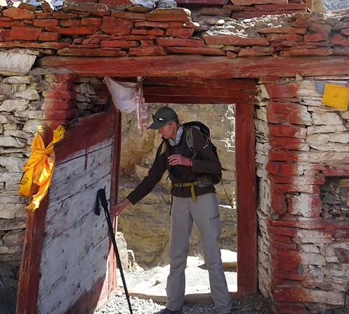 A hiker with a backpack and trekking pole passes through an ancient stone gateway decorated with colorful Buddhist prayer flags on the Nar Phu Valley Trek.