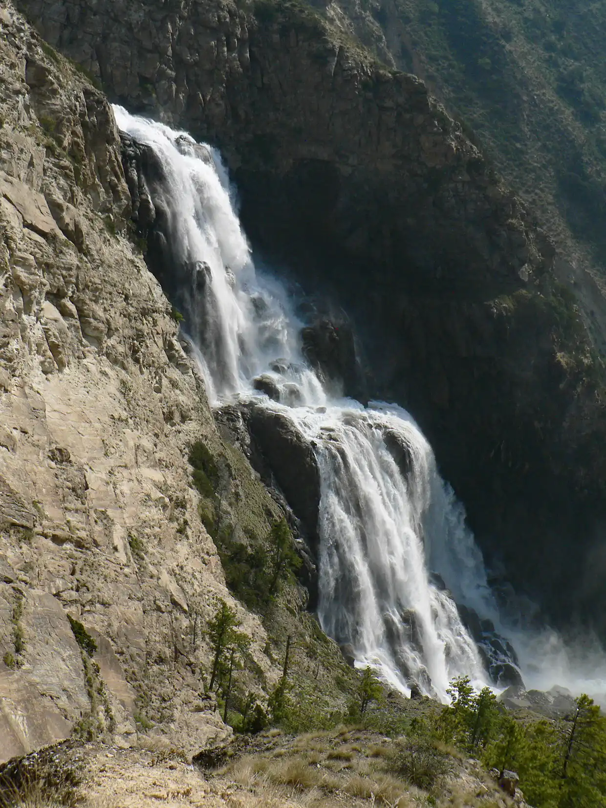 A powerful white waterfall cascades down a steep, rocky mountain cliff lined with sparse green trees during the scenic Upper Dolpo Trek.