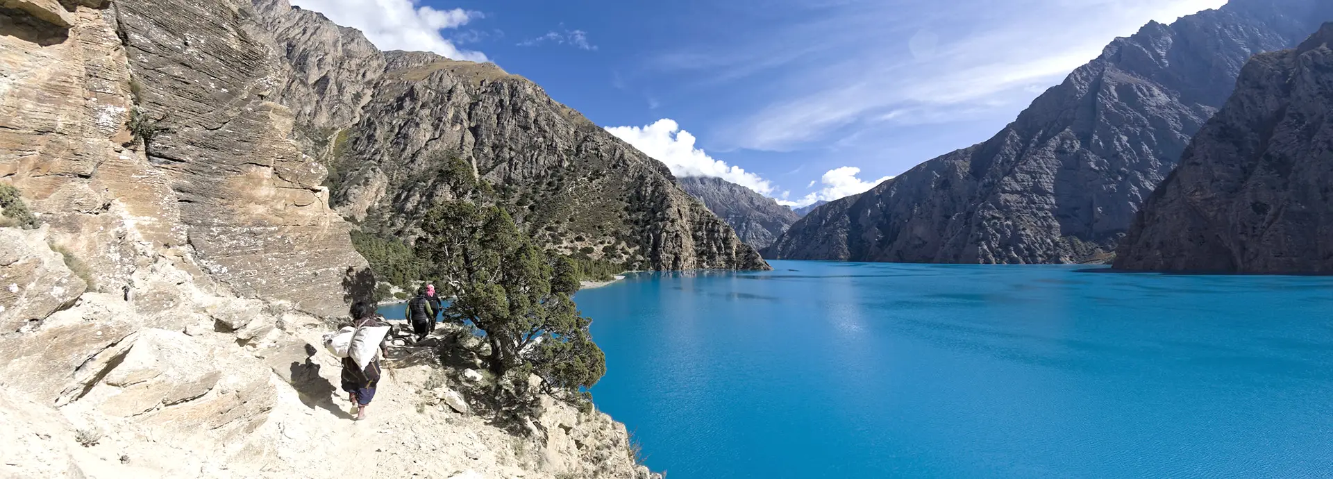 People navigating a rocky mountain trail alongside bright turquoise waters during the Shey Phoksundo Lake Trek, surrounded by steep cliffs and a clear blue sky.