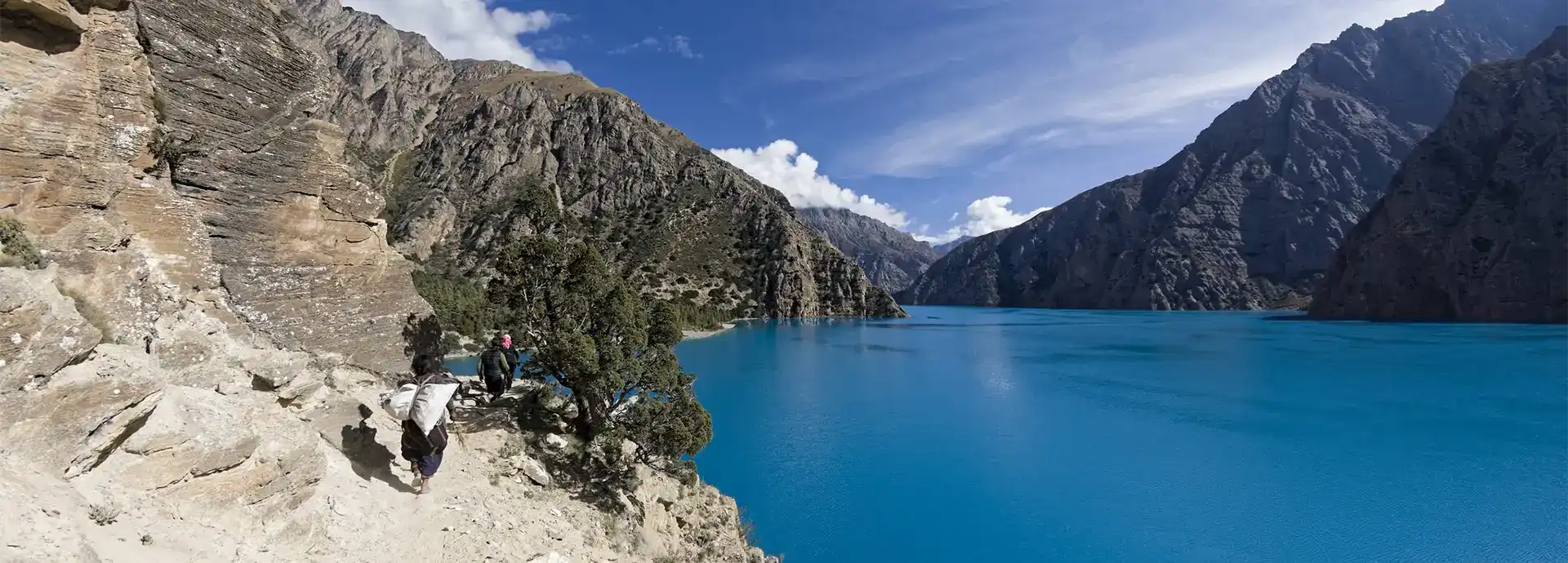 Trekkers walking along a narrow cliffside trail above the vibrant turquoise waters during the Shey Phoksundo Lake Trek